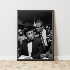 May include: Black and white photograph of Muhammad Ali and Malcolm X. Ali is wearing a tuxedo and is looking to the right. Malcolm X is wearing a suit and tie and is looking at Ali. The photo was taken in a crowded room.