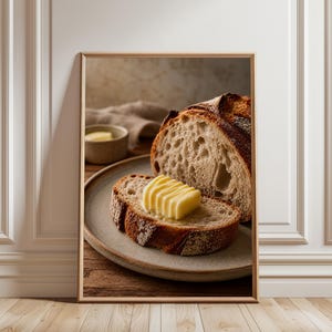 Puede incluir: Una impresión artística enmarcada que muestra una hogaza de pan de masa madre rústica cortada en un plato de cerámica. Una rebanada de pan está cubierta con una porción rectangular de mantequilla amarilla con estrías, junto a un cuenco pequeño.