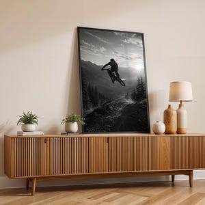 May include: A black and white framed wall art print depicting a mountain biker mid-air during a jump, set against a backdrop of mountains and pine trees. The artwork is displayed in a thin black frame, leaning against a light-colored wall.