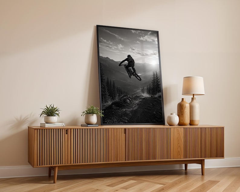 May include: A black and white framed wall art print depicting a mountain biker mid-air during a jump, set against a backdrop of mountains and pine trees. The artwork is displayed in a thin black frame, leaning against a light-colored wall.