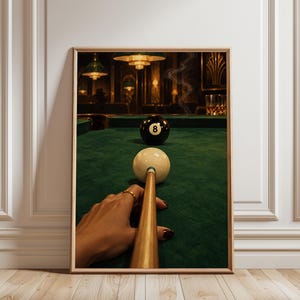 May include: A framed wall art print depicting a first-person perspective of a pool table with a green felt surface. A wooden pool cue rests against a white cue ball, aimed at a black eight ball. The background shows a dimly lit room with a glass.