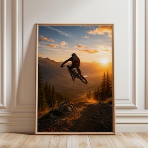 May include: A framed wall art print featuring a mountain biker mid-air during a jump against a sunset sky. The scene displays a silhouette of the rider and bike above a rugged mountain trail, surrounded by pine trees and distant peaks.