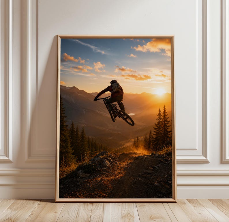 May include: A framed wall art print featuring a mountain biker mid-air during a jump against a sunset sky. The scene displays a silhouette of the rider and bike above a rugged mountain trail, surrounded by pine trees and distant peaks.