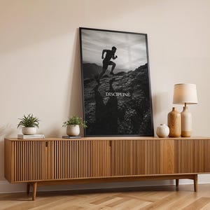 May include: A black and white framed wall art print featuring a silhouette of a person running uphill on a rocky mountain path. The word DISCIPLINE is printed in white capital letters in the center. The print is displayed in a thin black frame.