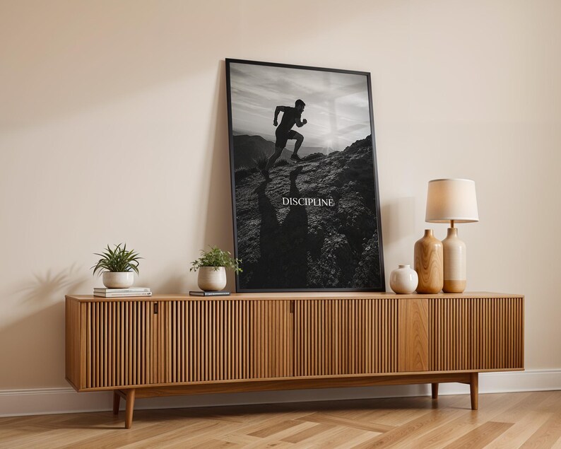 May include: A black and white framed wall art print featuring a silhouette of a person running uphill on a rocky mountain path. The word DISCIPLINE is printed in white capital letters in the center. The print is displayed in a thin black frame.