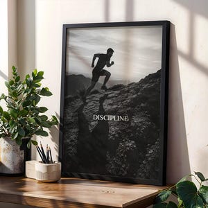 May include: A black and white framed wall art print featuring a silhouette of a person running up a rocky hill. The word DISCIPLINE is printed in white capital letters in the center of the image. The frame is black and rectangular in shape.