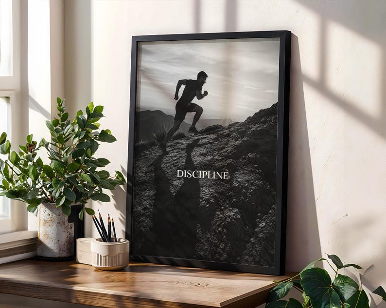 May include: A black and white framed wall art print featuring a silhouette of a person running up a rocky hill. The word DISCIPLINE is printed in white capital letters in the center of the image. The frame is black and rectangular in shape.