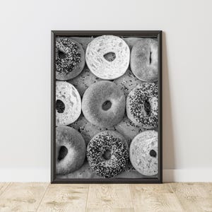 May include: Black and white framed print featuring nine bagels. Some bagels are covered in seeds or toppings, while others are plain. The bagels are arranged on a light-colored surface, creating a visually appealing food-themed artwork.