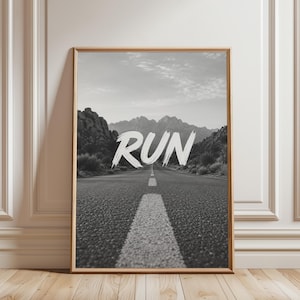 May include: A rectangular framed wall art print featuring a black and white landscape photograph of a long asphalt road stretching towards distant mountains, with the word RUN written in large, white, brush-stroke style lettering in the centre.