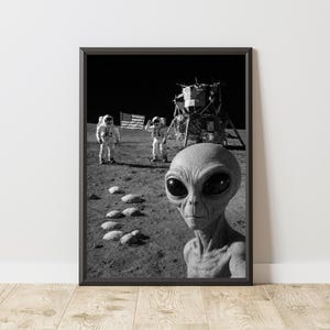 Op de afbeelding: Zwart-wit ingelijste print met een alien die een selfie maakt op de maan, met twee astronauten en de Amerikaanse vlag. De maanlander is zichtbaar op de achtergrond. De alien heeft grote zwarte ogen en een grijze teint.