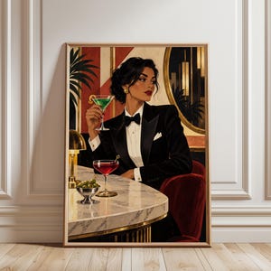 Op de afbeelding: Een ingelijste kunstdruk van een persoon met donker haar in een zwart smokingjasje, wit overhemd en zwarte vlinderdas, zittend aan een marmeren bar. De persoon houdt een groene cocktail vast, met een rode cocktail en een schaaltje olijven ernaast.