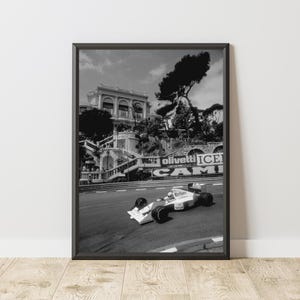 May include: A black and white photograph of a Formula 1 race car driving on a winding road. The car is a white and blue McLaren with the Marlboro logo on the side. The car is driving up a hill with buildings and trees in the background. The text "Olivetti ICE CAMI" is visible on a sign in the background.