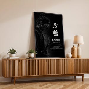 Op de afbeelding: Een zwart ingelijste verticale wandprint met een monochroom portret van een samoerai met een zwaard, de Japanse kanji voor Kaizen en Engelse tekst die de filosofie van constante verbetering uitlegt op een donkere achtergrond.