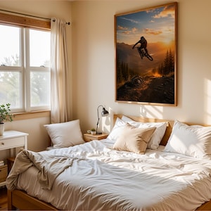 May include: A rectangular wall art print featuring a silhouette of a mountain biker jumping against a sunset sky over a forest, displayed in a bedroom with a wooden bed frame, white bedding, a side table with a lamp, and a window with curtains.