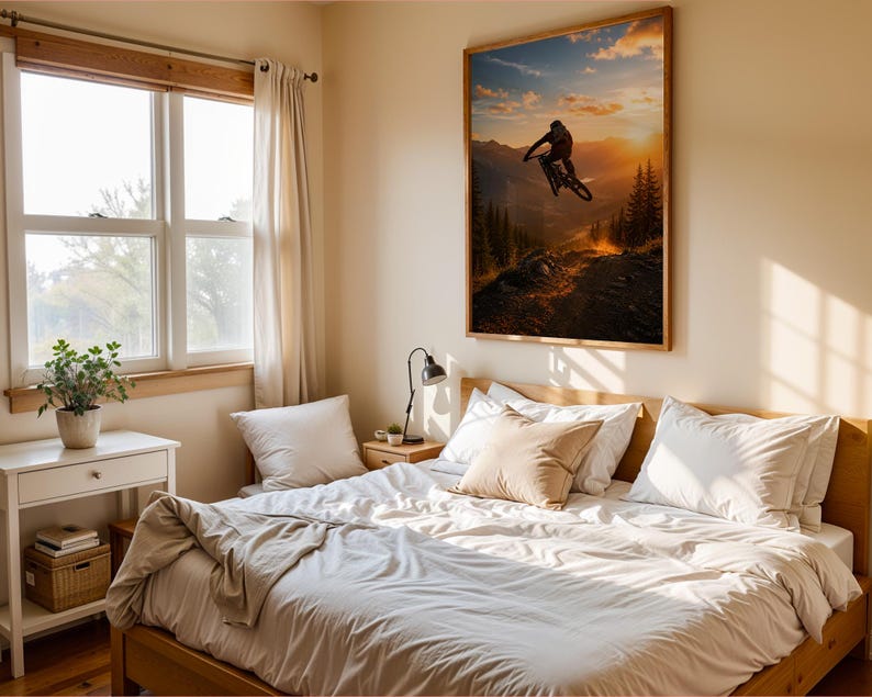 May include: A rectangular wall art print featuring a silhouette of a mountain biker jumping against a sunset sky over a forest, displayed in a bedroom with a wooden bed frame, white bedding, a side table with a lamp, and a window with curtains.