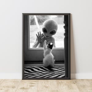 May include: Black and white framed print featuring an alien and a cat. The alien is behind a glass door, waving at a cat that is reaching up with a paw. The cat is black and white, and the alien has large black eyes.