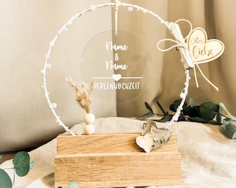 May include: A circular white wire hoop decorated with small white beads, mounted on a rectangular wooden base. A clear acrylic disc hangs in the center with the text Name & Name and PERLENHOCHZEIT. A wooden heart tag reads Alles Liebe.