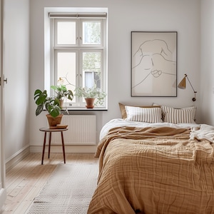 May include: A bedroom with a bed made with a brown quilted blanket and white and brown striped pillows. A black framed print of a line drawing of a person is hanging on the wall above the bed. A potted plant is on the windowsill and a small wooden side table with a potted plant is next to the bed.