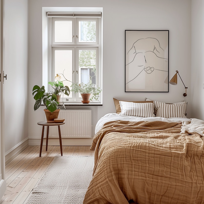 May include: A bedroom with a bed made with a brown quilted blanket and white and brown striped pillows. A black framed print of a line drawing of a person is hanging on the wall above the bed. A potted plant is on the windowsill and a small wooden side table with a potted plant is next to the bed.