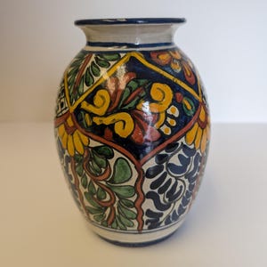 May include: A ceramic vase with a rounded body and a flared rim, featuring a hand-painted pattern of geometric diamonds filled with yellow, green, orange, and dark blue floral and leaf motifs against a cream-colored background with blue trim.