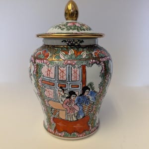 May include: A porcelain ginger jar with a lid featuring a gold-tone finial. The white surface is decorated with colorful hand-painted scenes of figures in traditional clothing, floral motifs, and geometric patterns in shades of pink, green, and orange.