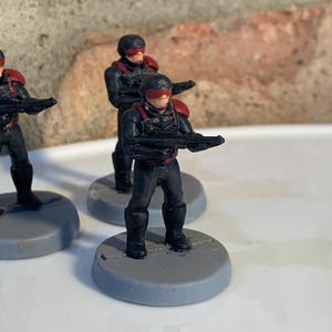 May include: Three black and red painted miniature figures of soldiers holding guns, standing on gray circular bases.