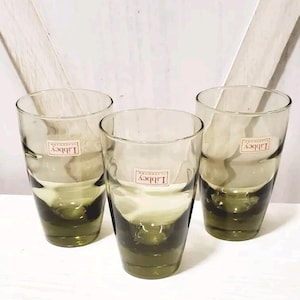 May include: Three clear drinking glasses with a green tint at the base. Each glass has a cylindrical shape and a label that reads "Libbey Glassware". The glasses are arranged on a light-colored surface.