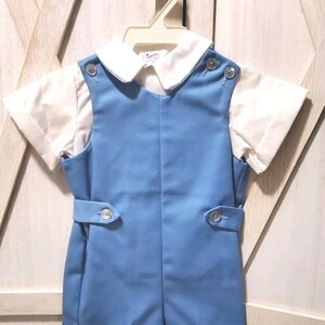 Puede incluir: Un conjunto de bebé azul claro con una camisa de cuello blanco y un peto. El peto tiene detalles de botones y una característica similar a un cinturón en la cintura. El conjunto está colgado en una percha blanca.