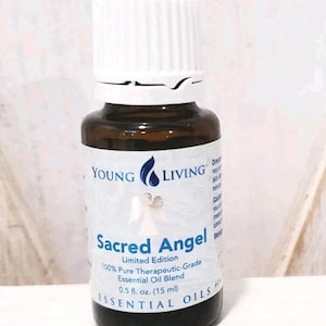 Może przedstawiać: Brązowa szklana butelka olejku eterycznego Sacred Angel firmy Young Living. Butelka ma białą etykietę z nazwą produktu i szczegółami. Butelka jest zwieńczona białą nakrętką. Etykieta wskazuje, że butelka zawiera 15 ml olejku eterycznego.