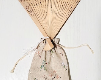 May include: A natural wood folding fan with intricate cut-out designs, paired with a beige drawstring pouch adorned with floral patterns in shades of green, pink, and white. The fan is open, and the pouch is closed.