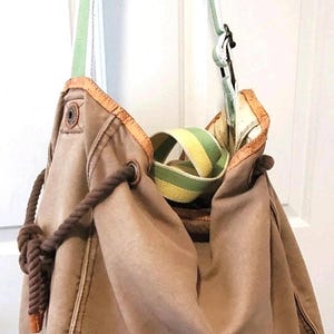 May include: A tan canvas hobo bag with a brown rope drawstring and a green and white striped strap. The bag has a brown trim and a yellow and green striped accent inside. A metal clasp secures the strap.