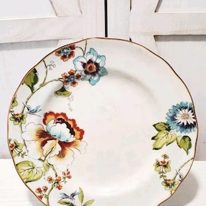 Puede incluir: Plato de cerámica blanca con un diseño floral que presenta grandes flores en tonos naranja, azul y amarillo, acentuadas con hojas verdes y un borde marrón. El plato se exhibe sobre un soporte de metal negro, mostrando un artículo de vajilla decorativo.