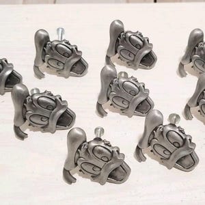 Puede incluir: Un conjunto de nueve pomos de armario de metal en tono plateado, cada uno con forma de cabeza de pato de dibujos animados con un pico y una gorra prominentes. Cada pomo tiene un tornillo de metal roscado en la parte posterior para fijar.