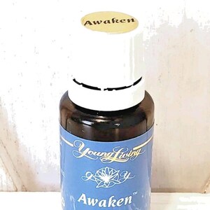 Op de afbeelding: Een bruine glazen fles Young Living Awaken essentiële olie. De fles heeft een witte dop en een blauw etiket met de productnaam en andere details. De fles bevat 15 ml essentiële oliën.