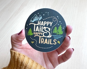 May include: A black round sticker with a white outline. The sticker has a gold and white design with the text "Happy Tails Happy Trails" and a mountain and tree graphic.