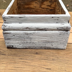 Puede incluir: Caja de madera cuadrada con un acabado de pintura blanca envejecida. La caja tiene un interior de madera natural y está sobre una superficie de madera. La caja probablemente se utiliza para almacenamiento o exhibición.