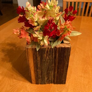 Puede incluir: Un jarrón de bloque de madera rústico lleno de flores artificiales coloridas. Las flores son una mezcla de tonos rojos, melocotón y crema, con hojas verdes. El jarrón está hecho de madera envejecida y texturizada.