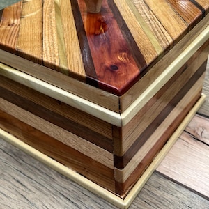 Puede incluir: Una caja de madera hecha a mano con tapa. La caja está construida con varios tipos de madera en tonos marrón, beige y rojo, dispuestos en un patrón de rayas. Un pequeño pomo de aspecto natural sirve de asa en la tapa. La caja está sobre una superficie de madera.