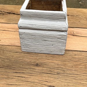 Puede incluir: Una jardinera cuadrada de madera con un acabado pintado de blanco envejecido. La caja tiene un diseño en capas y se apoya sobre una superficie de madera. El interior de la caja es de madera natural.