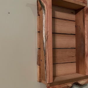 May include: A rustic wooden wall shelf with a natural edge design. The shelf is constructed from light brown wood with visible grain and a rough, unfinished edge. The interior features horizontal wooden slats.