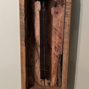 May include: A rustic, framed artwork featuring a dark, weathered metal object, possibly a chisel or similar tool. The object is set against a backdrop of textured, reddish-brown wood, encased within a rough-hewn wooden frame.