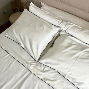 Pode incluir: Lençóis brancos para cama com acabamento preto. Os lençóis estão bem feitos e a cama está arrumada.