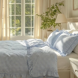 Könnte beinhalten: Ein hellblau-weiß karierter Bettbezug und passende Kissenbezüge mit Rüschenkanten. Das Bettwäscheset ist auf einem Bett mit einem weißen Spannbettlaken arrangiert, in einem Raum mit cremefarbenen Vorhängen und einem Fenster.