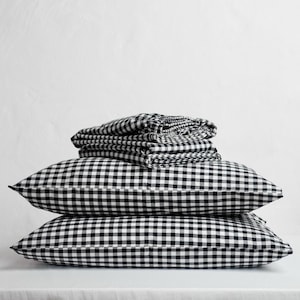 May include: A set of black and white gingham bedding, comprising two pillows and folded sheets. The pillows are stacked, with the sheets neatly folded on top. The bedding is arranged on a white surface, creating a clean, minimalist aesthetic.