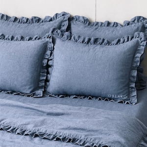 May include: A set of blue bedding with ruffled edges. The set includes pillowcases and a duvet cover. The pillowcases have the word "OBLAKO" embroidered on them. The bedding is made of a soft, textured fabric.