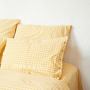 May include: A set of yellow and white gingham bedding, including pillowcases and a fitted sheet. The pillowcases have button closures. The word "OBLAKO" is visible on one of the pillowcases. The bedding is arranged on a bed against a white wall.