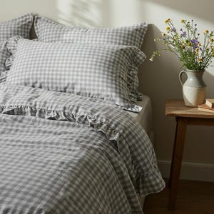 May include: A gray and white gingham patterned bedding set. The set includes pillowcases and a duvet cover with ruffled edges. A small wooden table holds a vase of flowers.