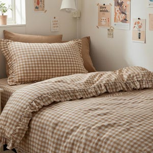 May include: A bed set featuring a tan and white gingham pattern. The set includes a pillowcase with a ruffled edge, a fitted sheet, and a duvet cover. The bedding is arranged on a bed with a neutral-toned headboard and wall.