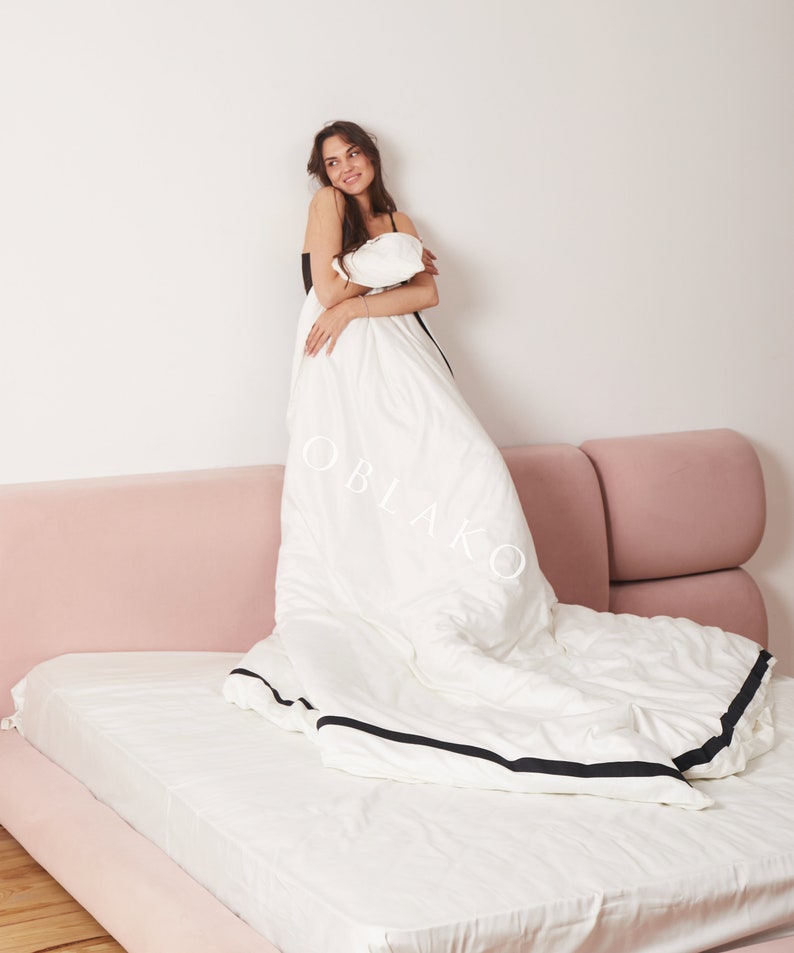 Pode incluir: Uma capa de edredom branca com uma borda preta, deitada em uma cama rosa. Uma pessoa est&aacute; em p&eacute; na cama, segurando a capa de edredom.