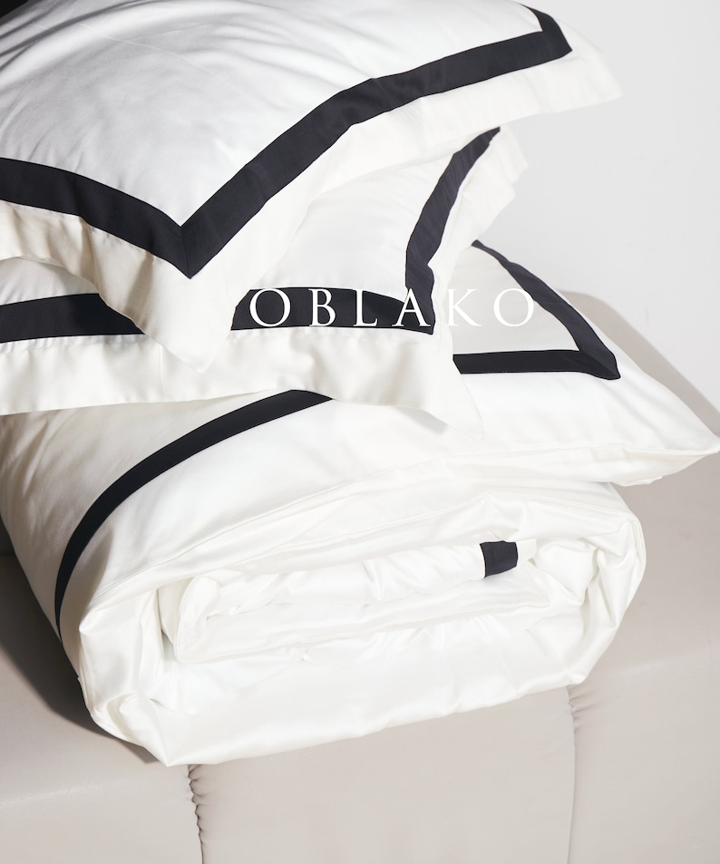 Pode incluir: Roupa de cama branca com acabamento preto. A roupa de cama est&aacute; dobrada e empilhada, com a camada superior dobrada para revelar o acabamento preto. A roupa de cama est&aacute; etiquetada como "OBLAKO".
